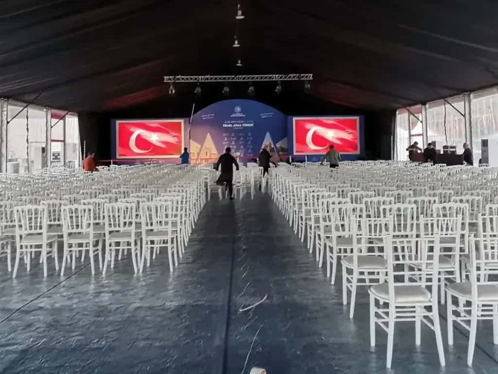 Çadır içinde tören düzeni: sahne, iki LED ekran ve yüzlerce beyaz sandalye